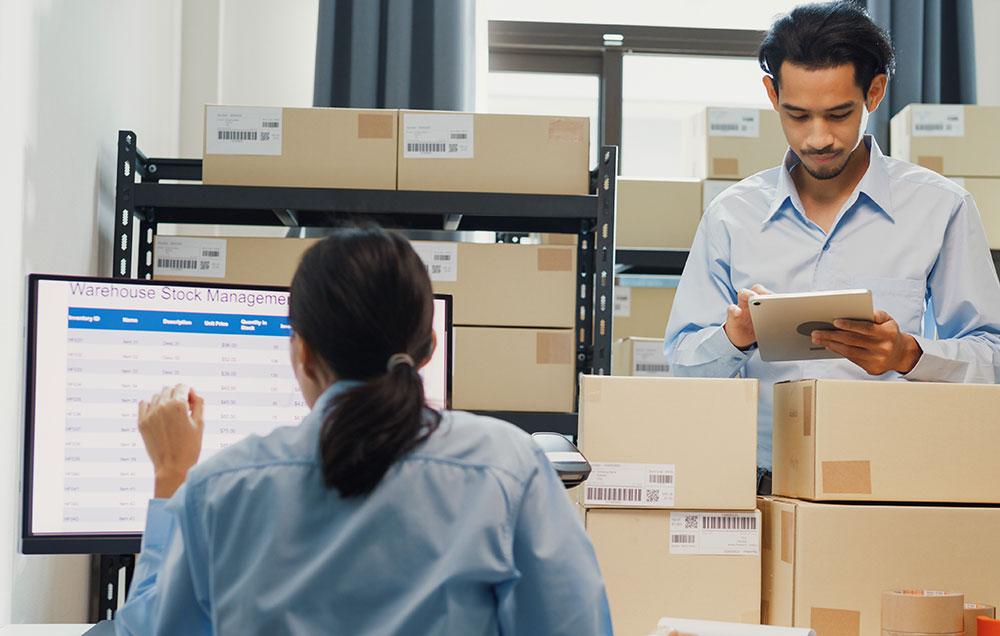 Photograph of two people in a stock room; one takes notes on a tablet while the other has a stock management system open on a computer.