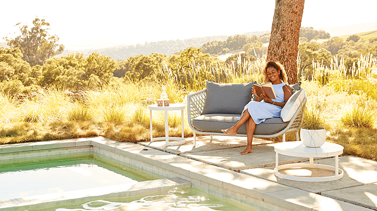 A smiling young woman reading a book while sitting on stylish patio furniture next to a pool.